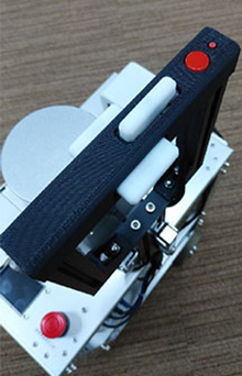 清水建設版エーアイスーツケース五号機のハンドルの写真。ハンドル上には前方に操作のための大きいボタンが一つ、右側面に小さいボタンが一つ、ついています。ハンドルの上の面と左右の側面にある振動ロータは四号機と同様です